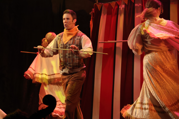 A performer in a waistcoat balances objects, surrounded by dancers in flowing costumes.