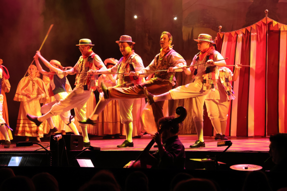 A group of performers in striped costumes dancing on stage with a vibrant backdrop.