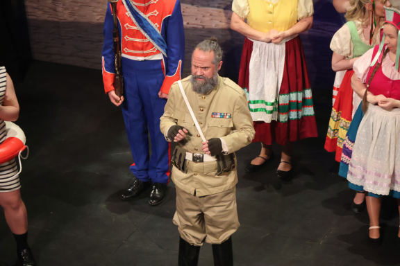 A man in a beige outfit stands centre stage, surrounded by performers in colourful costumes.