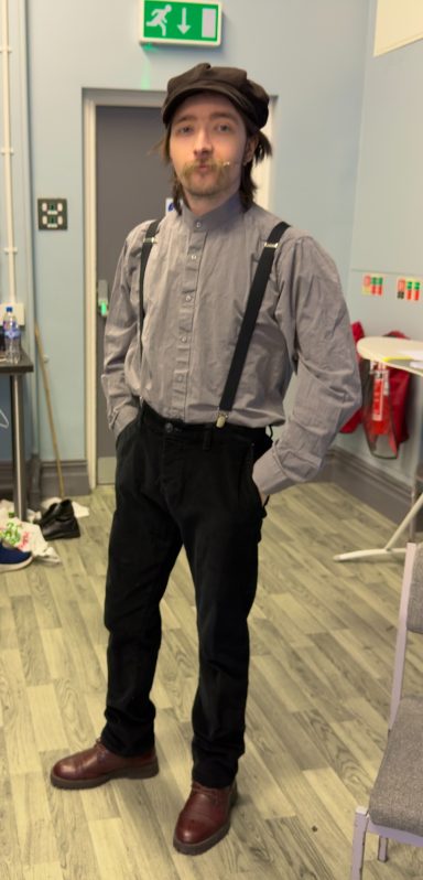 Person dressed in a vintage outfit with suspenders and brown boots, standing indoors.