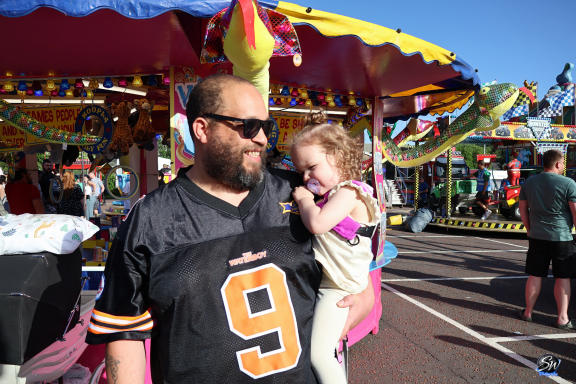 Man wearing sunglasses holds a small child at a carnival. Brightly coloured attractions in background.