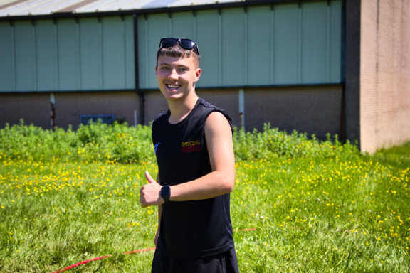 Young man in a sleeveless top smiles and gives a thumbs up in a grassy outdoor setting.