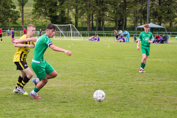 Two young football players compete for the ball on a grassy pitch, with spectators in the background.