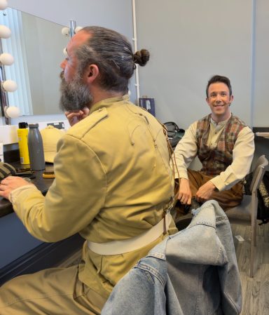 Two men in a dressing room, one seated and another adjusting his beard at a vanity.