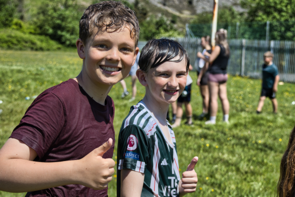 Two boys smiling and giving thumbs up, with a group playing in the background.