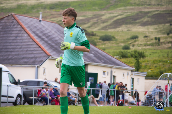A young goalkeeper in green kit stands on a football pitch with spectators in the background.