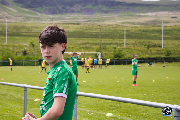 A young boy in a green football kit watches training on a grass pitch.