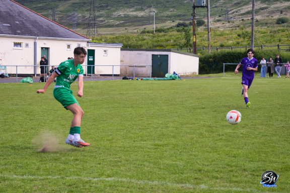 A footballer in green kicks a ball on a grassy pitch, with a goalkeeper in purple nearby.