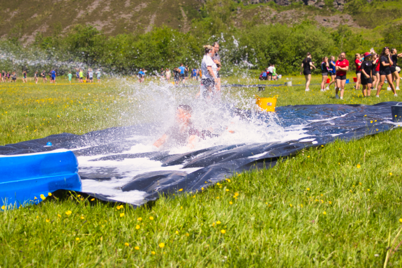 A person sliding down a water-covered mat on a grassy field, with spectators in the background.