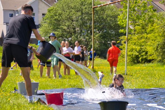 Child splashing in water as another person pours water from a bucket during a fun outdoor event.