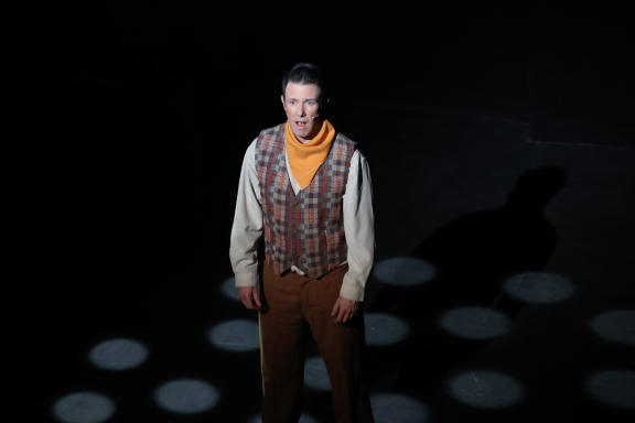 A man in period costume stands under spotlight, casting a shadow on the floor.