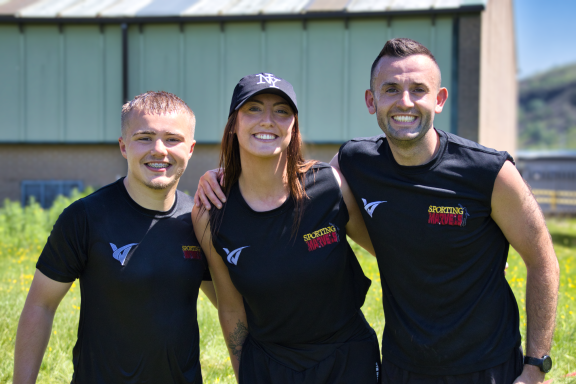 Three smiling individuals in black tank tops pose outdoors, with green grass and buildings behind.