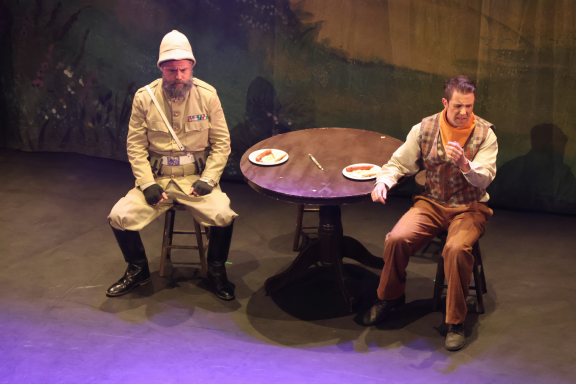 Two seated actors on stage, one in a white uniform and the other in brown, by a table.