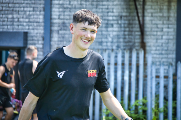 Young man smiling in a black t-shirt, with a blurred outdoor background.
