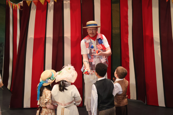A performer entertains children in period costumes on a stage with striped curtains.