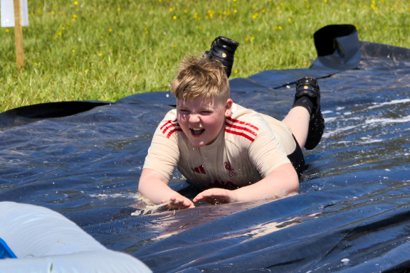 A child wearing a beige shirt slides joyfully on a black tarp in a grassy field.