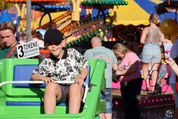 A smiling boy in a patterned shirt sits on a fairground ride, surrounded by festive activities.