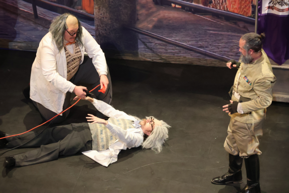 Two scientists in lab coats attend to a unconscious man on stage, while another stands nearby.