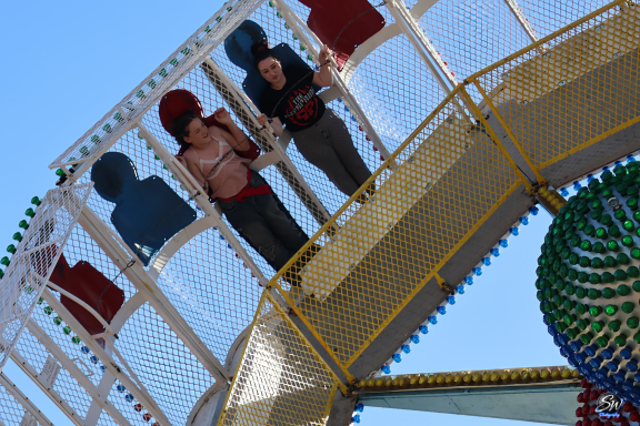 Two people are standing on a ferris wheel carriage against a blue sky.