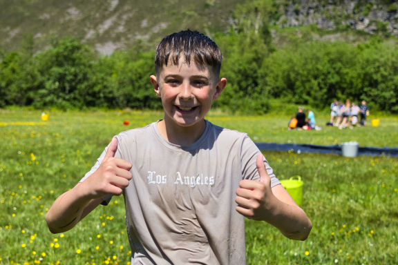 A smiling boy giving a thumbs-up in a grassy field with friends in the background.
