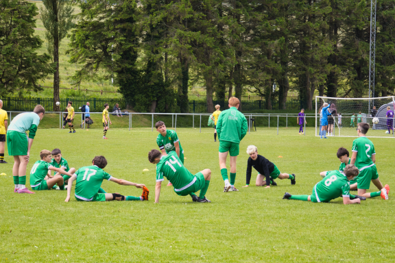 Football players in green kits stretching on a grassy field, surrounded by teammates.