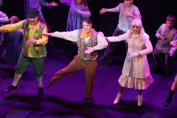 Three performers dancing on stage, wearing colourful costumes, in a lively musical scene.