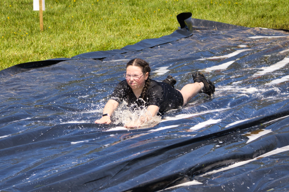 A person in a black outfit slides on a wet tarp, splashing water in a grassy area.