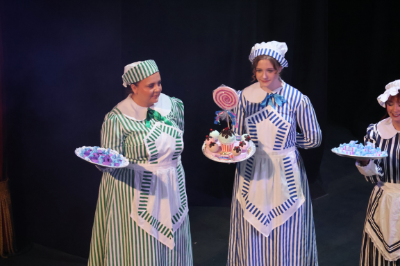 Three women in historical costumes hold colourful desserts on trays.