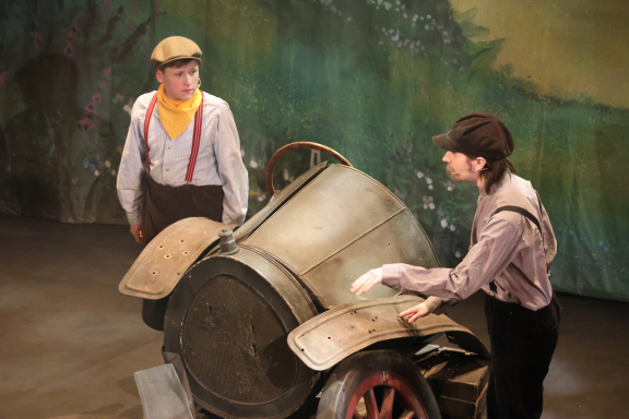 Two actors in vintage costumes interact with an old-fashioned vehicle on stage.