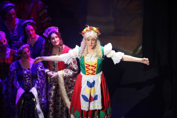 A performer in a colourful costume with arms outstretched, surrounded by other characters.