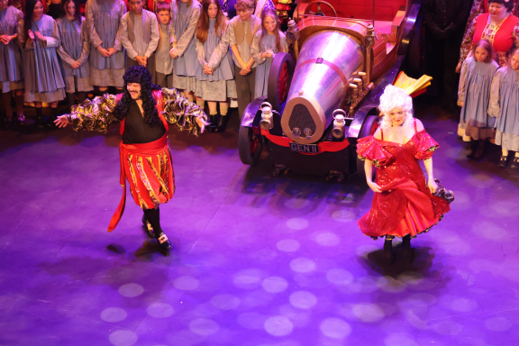Two performers in vibrant costumes dancing on stage with a crowd in the background.