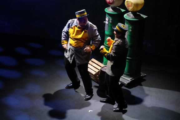 Two performers in costumes dancing on stage, with props in the background.