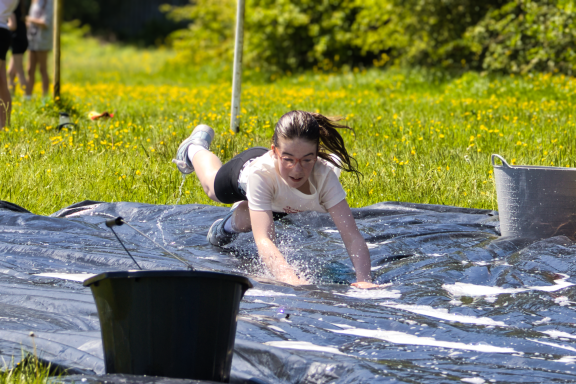 A girl in a white shirt slides on a water-covered tarp in a grassy field.