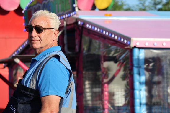 Man in sunglasses, wearing a blue shirt, standing near a colourful carnival attraction.